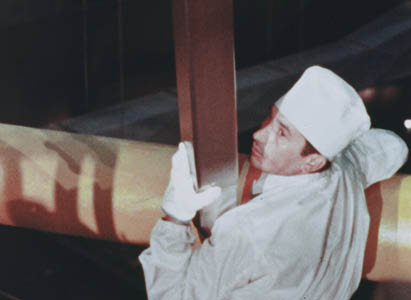 Man in white coveralls, a white cap, and white gloves holds a suspended fresh nuclear fuel assembly close while positioning it for initial core loading at BONUS.