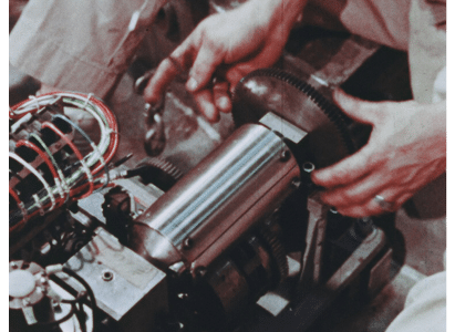 Control rod drive mechanism, which is a shiny steel cylinder with a larger gear sticking out the end of it. A mans two hands are seen nearly grasping the gear.