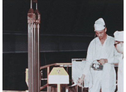 Men in white coveralls and caps look at geiger counter while standing near a nuclear superheat fuel assembly being loaded into BONUS. Dark background. Radiation sign on pedastal.