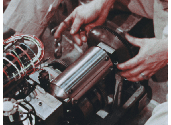 Control rod drive mechanism, which is a shiny steel cylinder with a larger gear sticking out the end of it. A mans two hands are seen nearly grasping the gear.