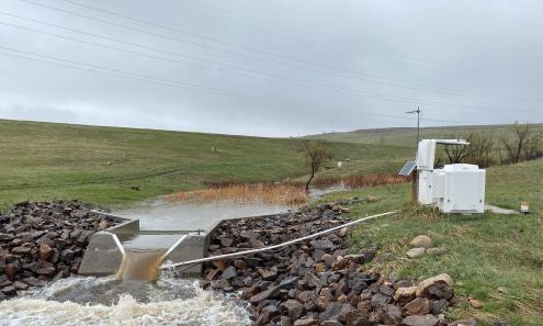 A concrete structure is located near a river, with a small pond nearby. The area is surrounded by rocks and grass, and there is a small building nearby. AI generated content
