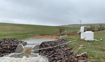A concrete structure is located near a river, with a small pond nearby. The area is surrounded by rocks and grass, and there is a small building nearby. AI generated content