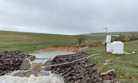 A concrete structure is located near a river, with a small pond nearby. The area is surrounded by rocks and grass, and there is a small building nearby. AI generated content